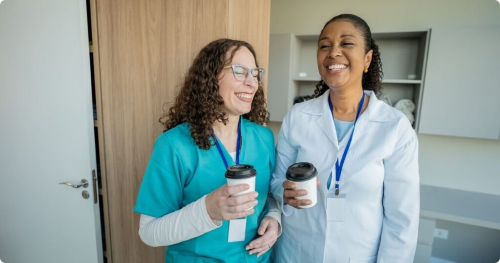 Two nurses standing together in a hospital break room, sharing a conversation, humor, and a moment of support.