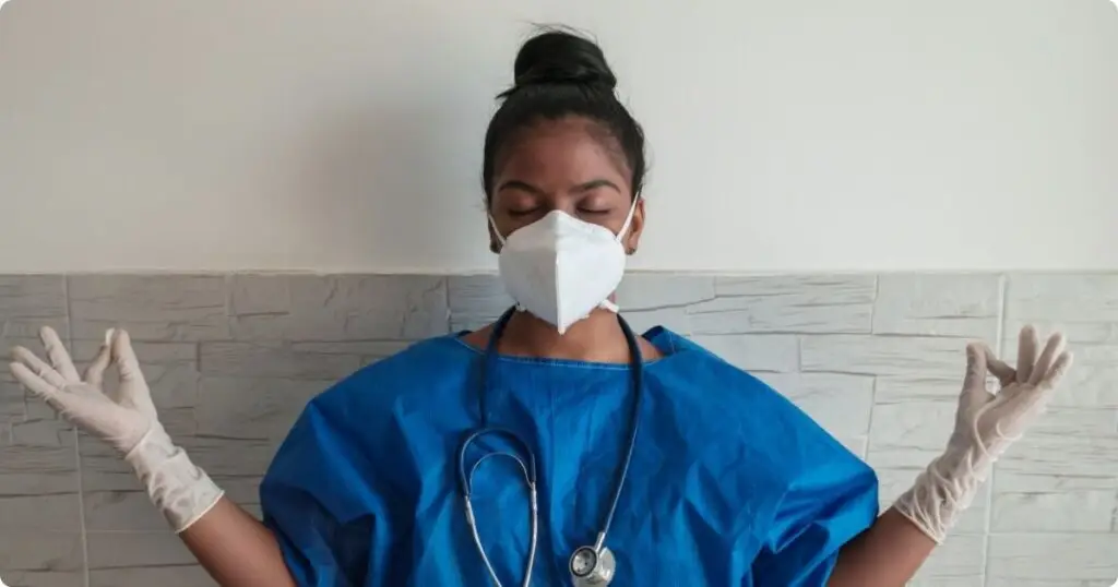 A nurse sits in a meditation pose in the hallway. She is taking a deep breath, representing self-care and emotional recovery from compassion fatigue.